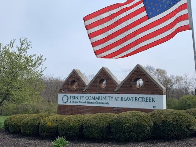 A brick sign for Trinity Community at Beavercreek with the text 'A United Church Homes Community' beneath the name. The sign is surrounded by neatly trimmed bushes and greenery. Above the sign, an American flag is prominently displayed on a flagpole. Trees and a cloudy sky are visible in the background.
