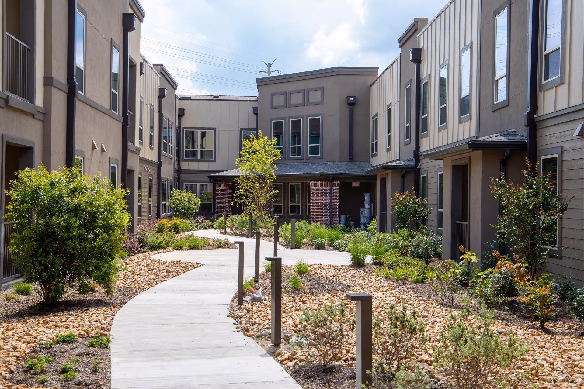 Outdoor courtyard area at Watercrest at Shadow Creek Ranch featuring a curved concrete walkway surrounded by landscaped garden beds with small trees, shrubs, and rocks, flanked by two-story residential buildings with multiple windows.