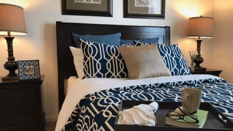A neatly made bed with navy-and-white patterned bedding, decorative pillows, a dark wood headboard, nightstands with lamps, and a tray holding a mug and glasses.