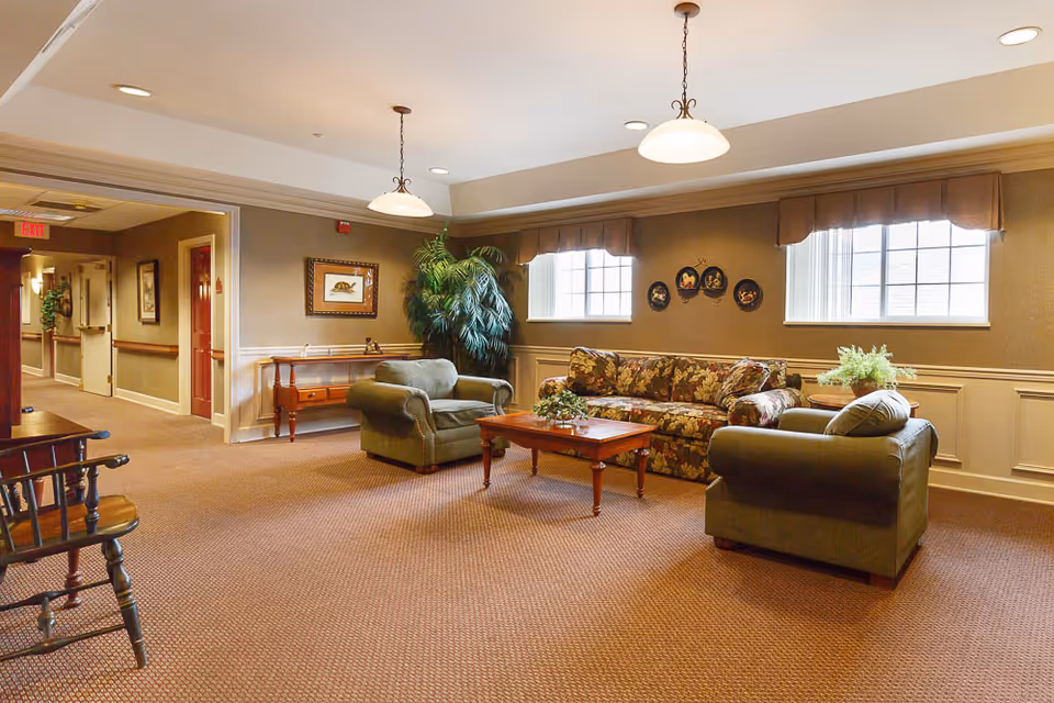 A cozy sitting area in a senior living facility with a floral-patterned sofa, two green armchairs, a wooden coffee table, and a side table with a plant. The room has beige walls with wainscoting, two windows with valances, framed artwork on the walls, and a large potted plant in the corner. The carpet is brown, and there are pendant lights hanging from the ceiling. A hallway with doors and an exit sign is visible in the background.