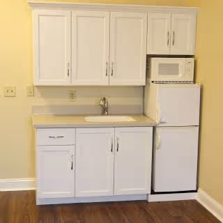 Small kitchenette with white upper and lower cabinets, a sink, microwave and compact refrigerator against a pale yellow wall.