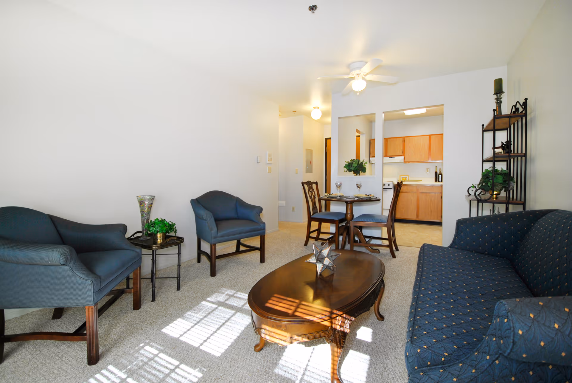 A bright living room area with a blue patterned sofa, two blue armchairs, a wooden coffee table with a decorative star-shaped object, and a small round dining table with two chairs set with wine glasses. The room has light-colored walls and carpet, a ceiling fan with a light, and an open view into a kitchen with wooden cabinets.