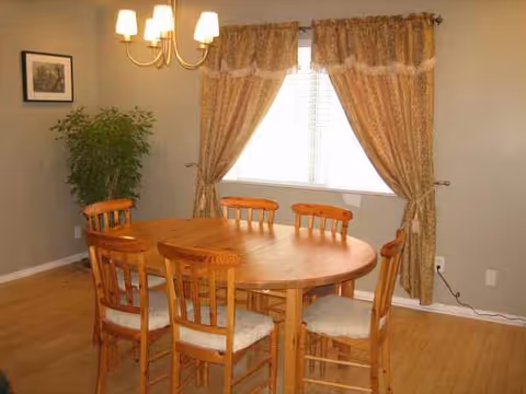 Round wooden dining table with six chairs, window with drapes, and a chandelier.
