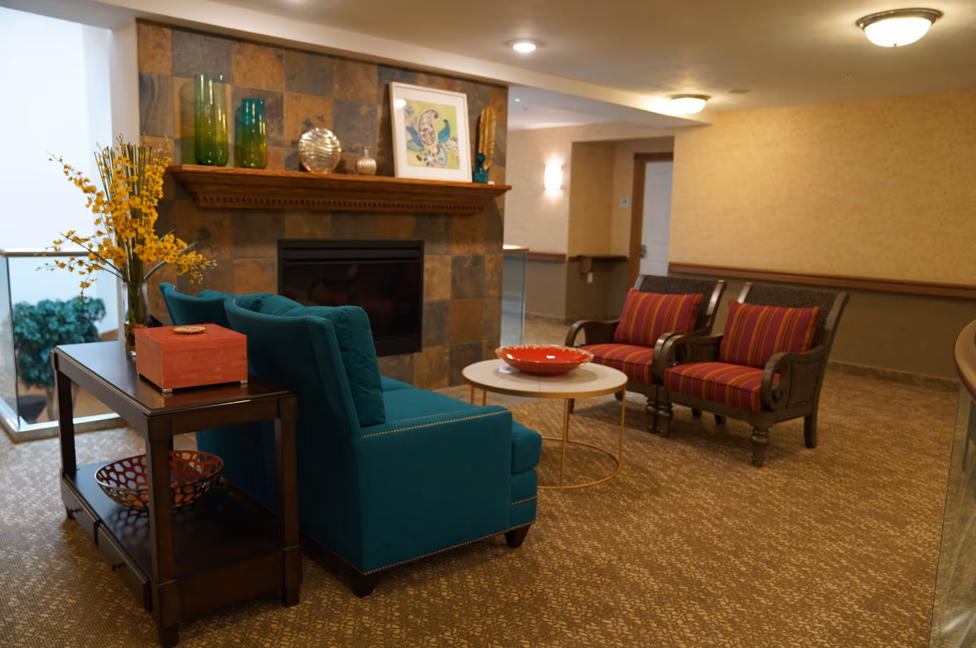 A cozy seating area in a retirement community featuring a teal upholstered sofa, two wooden armchairs with red and purple striped cushions, a round white coffee table with a red decorative bowl, and a fireplace with a stone tile surround. The room has warm lighting, a wooden side table with decorative items including a vase with yellow flowers, and a framed artwork on the mantel.