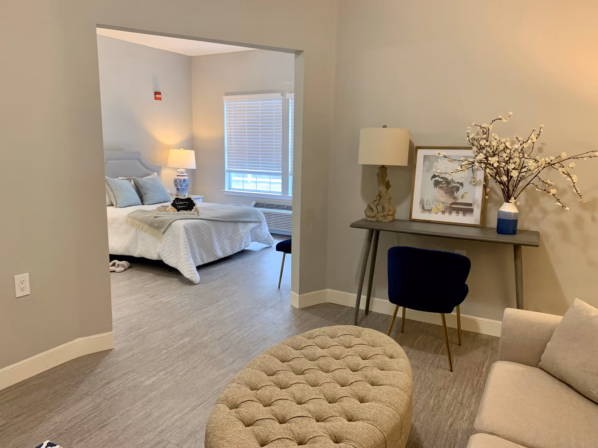 Open-plan senior apartment living area with a tufted ottoman and sofa, a small desk with lamp and vase of flowers, and a bed visible through an open doorway.