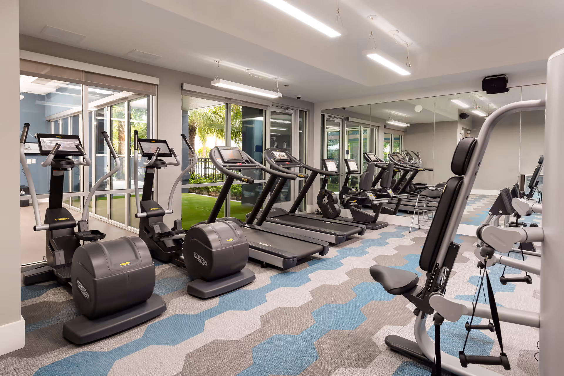 A modern fitness room with exercise equipment including treadmills, elliptical machines, and a weight machine. The room has large windows showing an outdoor garden area and a wall covered with mirrors. The floor has a patterned carpet in shades of blue, gray, and beige.
