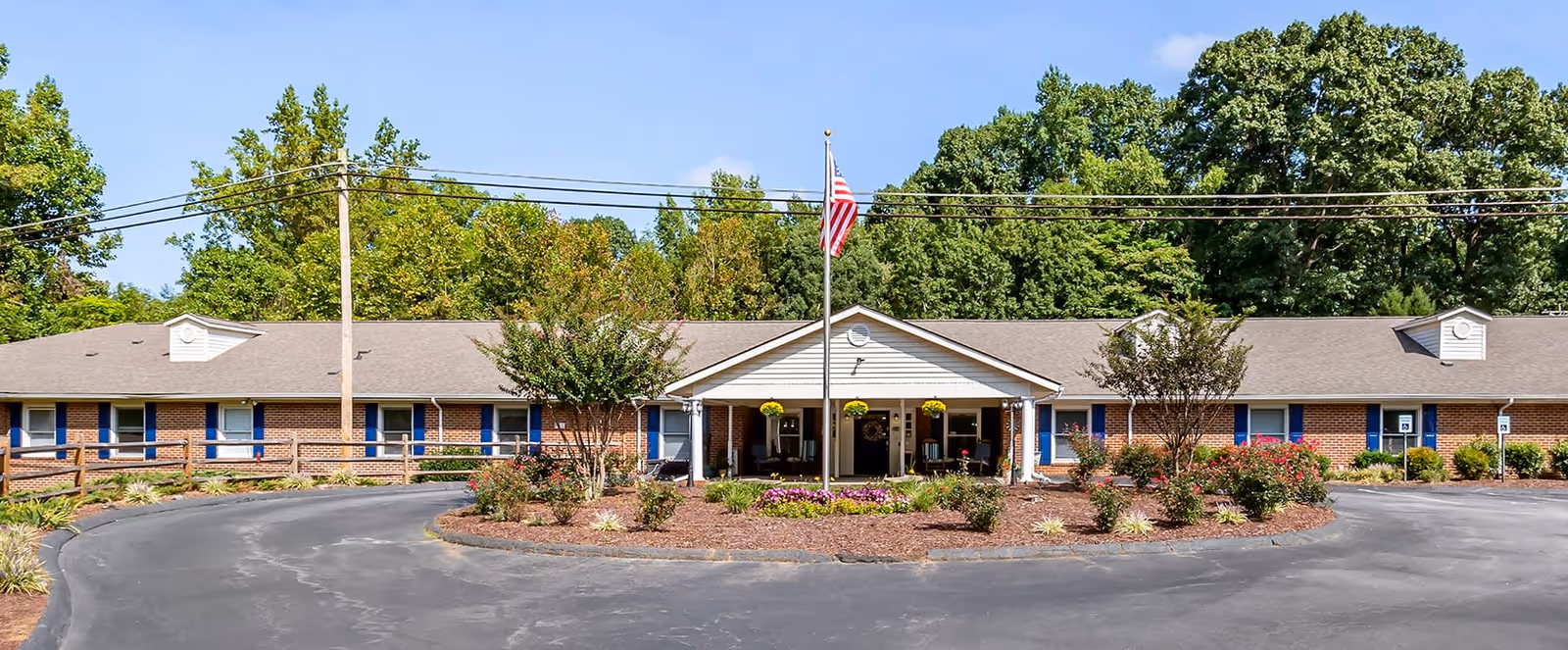 Front exterior view of Brookdale East Broad facility, a single-story brick building with a gray roof, blue window shutters, and a covered entrance with white columns. There is a circular driveway with landscaped flower beds and an American flag on a flagpole in front. Trees and greenery are visible in the background under a clear blue sky.
