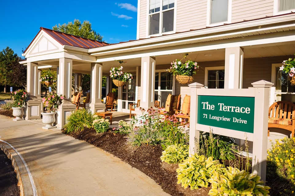 Front porch entrance of The Terrace senior living facility with a green sign reading 'The Terrace 71 Longview Drive', hanging baskets, chairs, and landscaping.