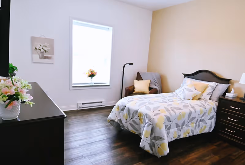 A well-lit bedroom with a bed, dresser, and flowers by a window.