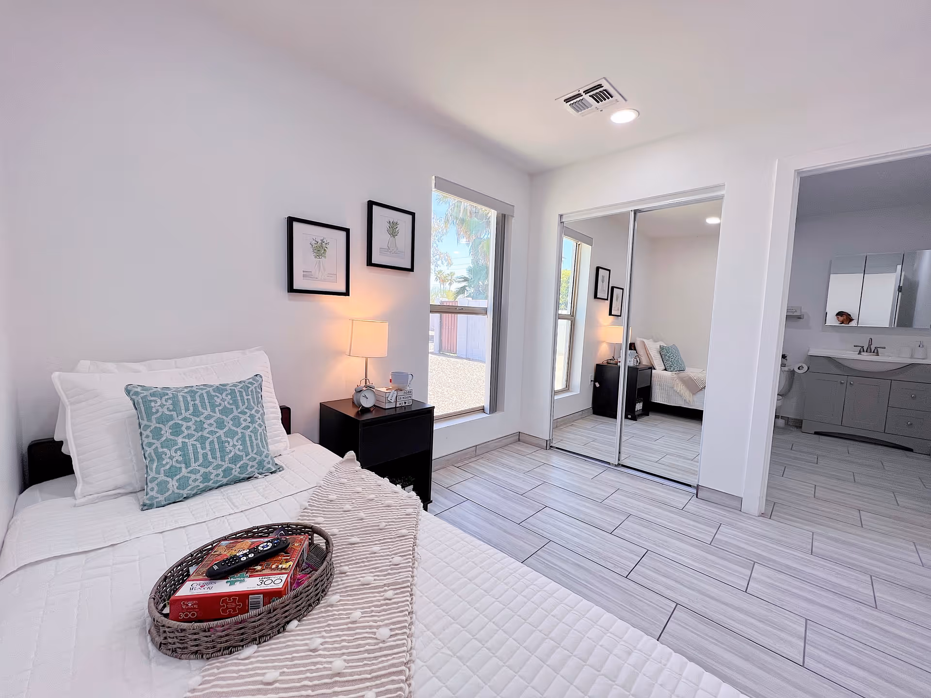 A bright and clean bedroom with a single bed covered in white bedding and a decorative blue pillow. A small black nightstand with a lamp, clock, and cup sits next to the bed. Two framed botanical prints hang on the wall above the nightstand. There is a large window letting in natural light and a mirrored closet door reflecting the room. The floor is tiled in a light gray color, and an open doorway leads to a bathroom with a gray vanity and mirror.