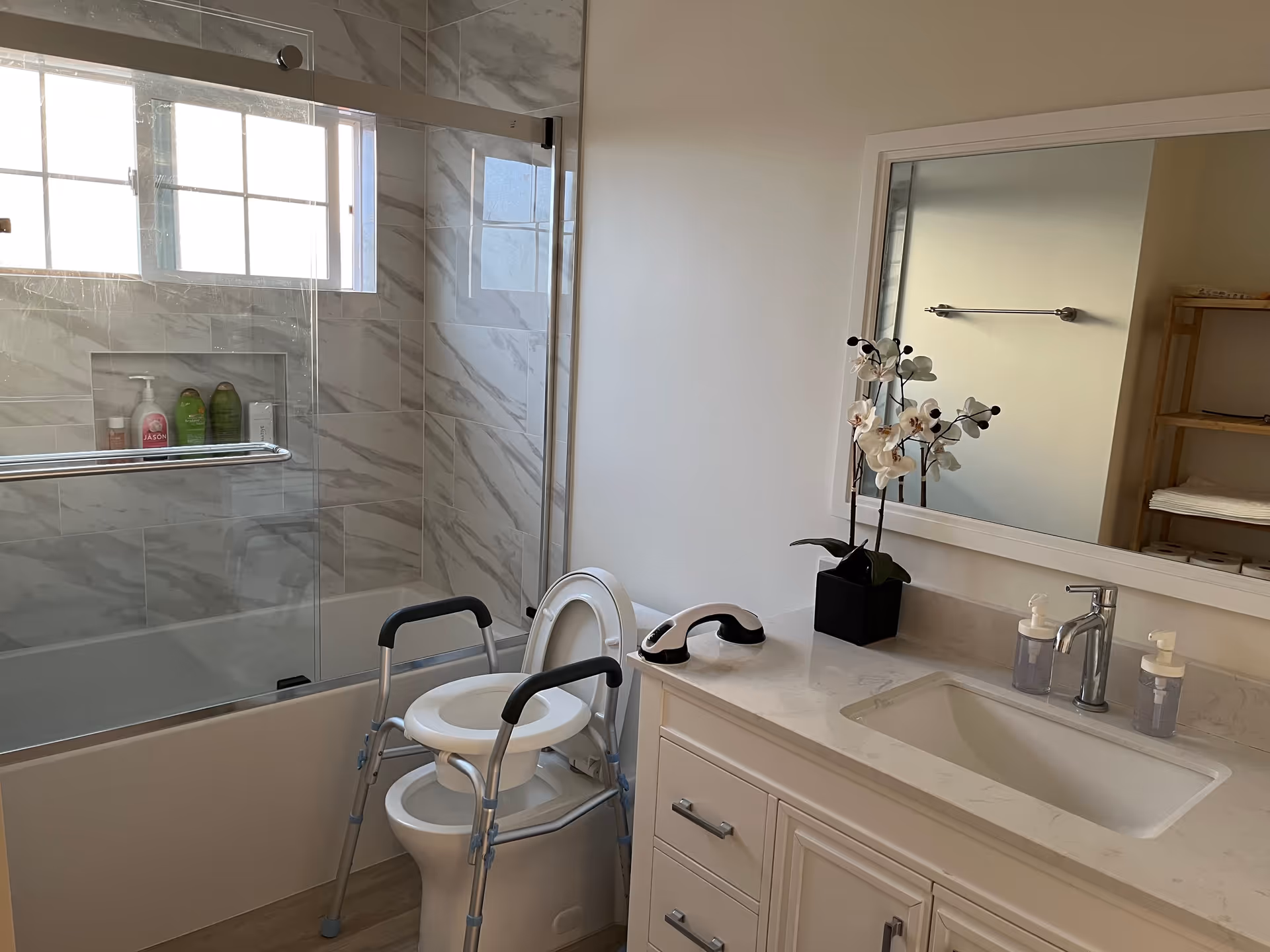 A clean and modern bathroom with a bathtub and shower combination featuring marble-patterned tiles and a glass sliding door. There is a white toilet with a raised toilet seat and safety frame for assistance. A white vanity with a sink, two soap dispensers, a black potted orchid plant, and a large mirror above it is visible. A wooden shelf with folded towels is reflected in the mirror.