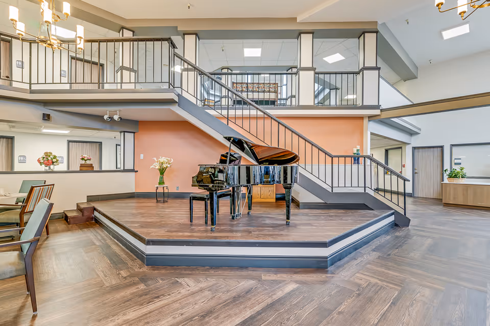 A spacious senior living facility common area featuring a black grand piano on a raised wooden platform beneath a staircase. The room has wood flooring, a peach-colored accent wall, and a railing along the upper level. There are chairs and tables to the left and some decorative flowers on the walls and tables.
