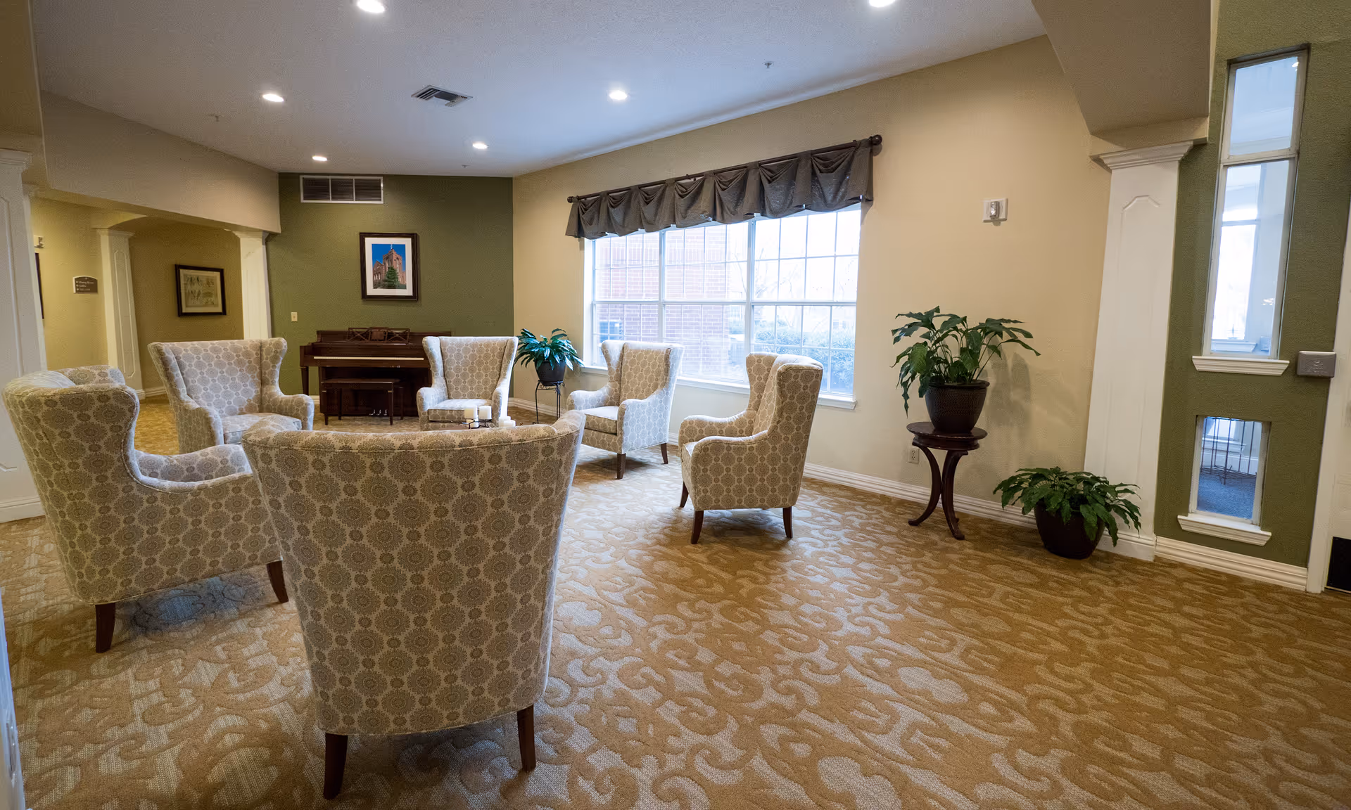 A bright lounge with patterned armchairs arranged around a coffee table, a piano against the far wall, plants, and a large window.