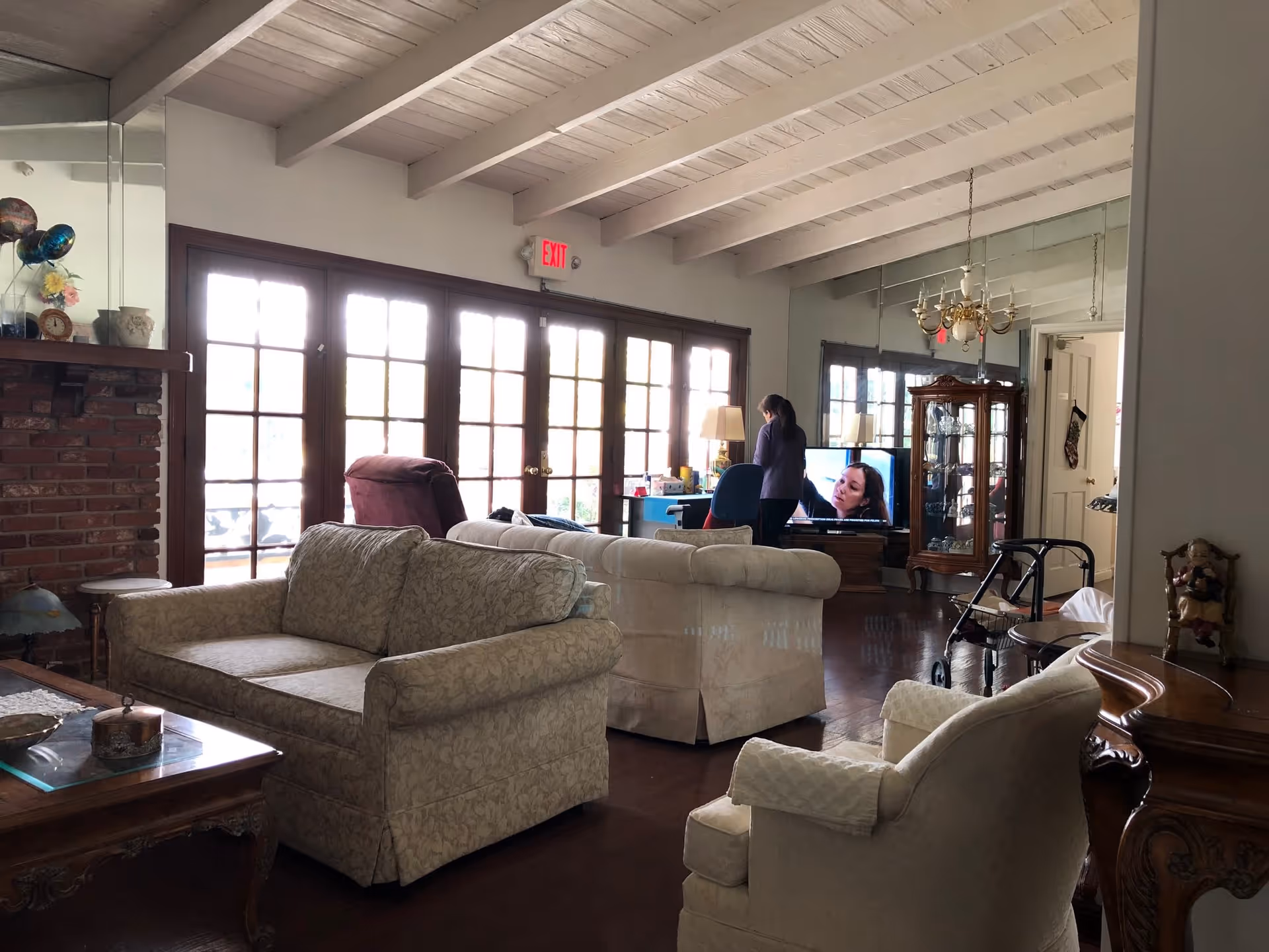 Living room with upholstered sofas and armchairs, wood-beamed ceiling, brick fireplace, French doors, and a TV with a person standing near a desk.