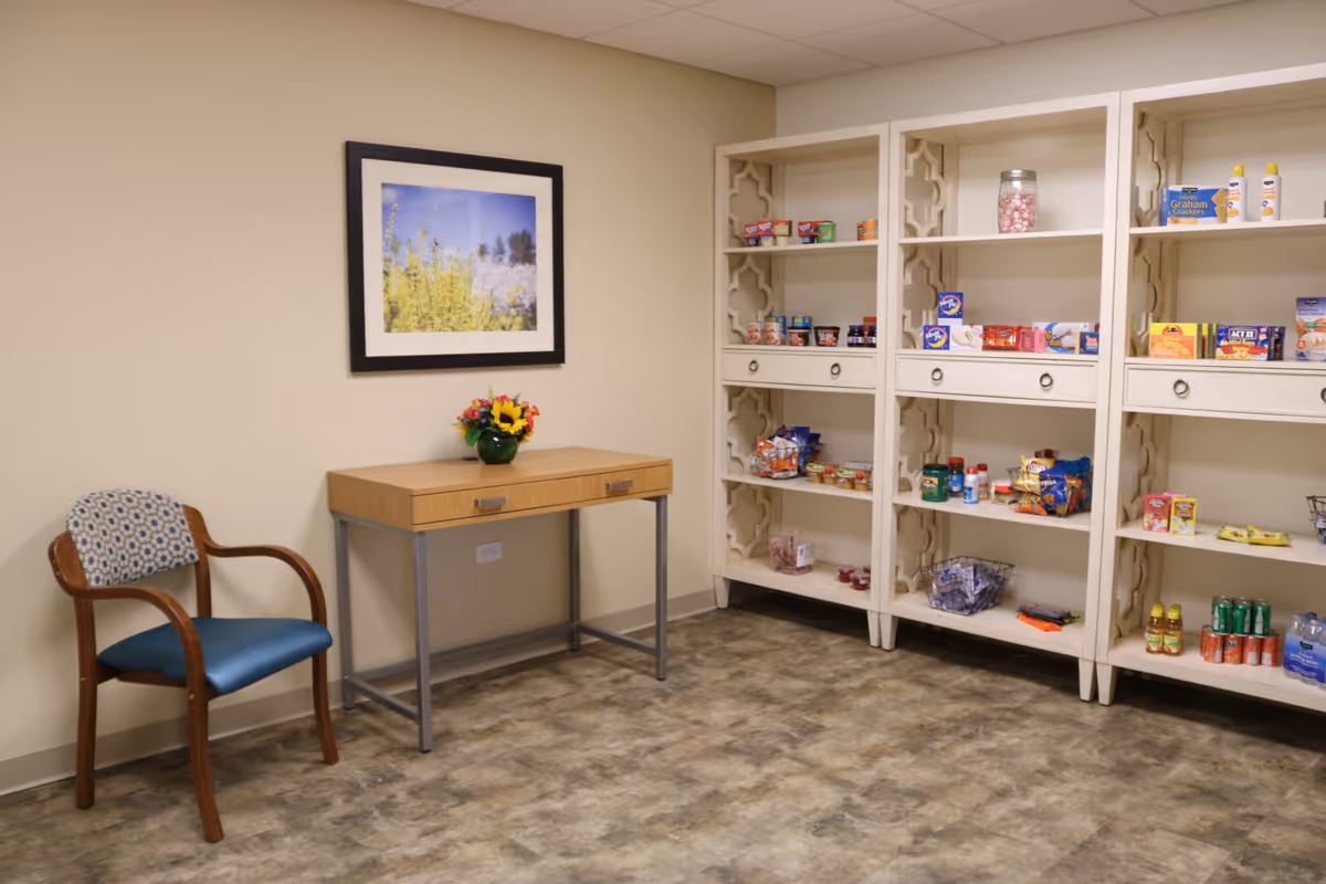 A small room with a wooden chair with blue cushion and patterned backrest, a wooden table with metal legs holding a small vase of colorful flowers, and white shelves stocked with various snacks and drinks. A framed picture of yellow flowers and a blue sky hangs on the beige wall above the table.