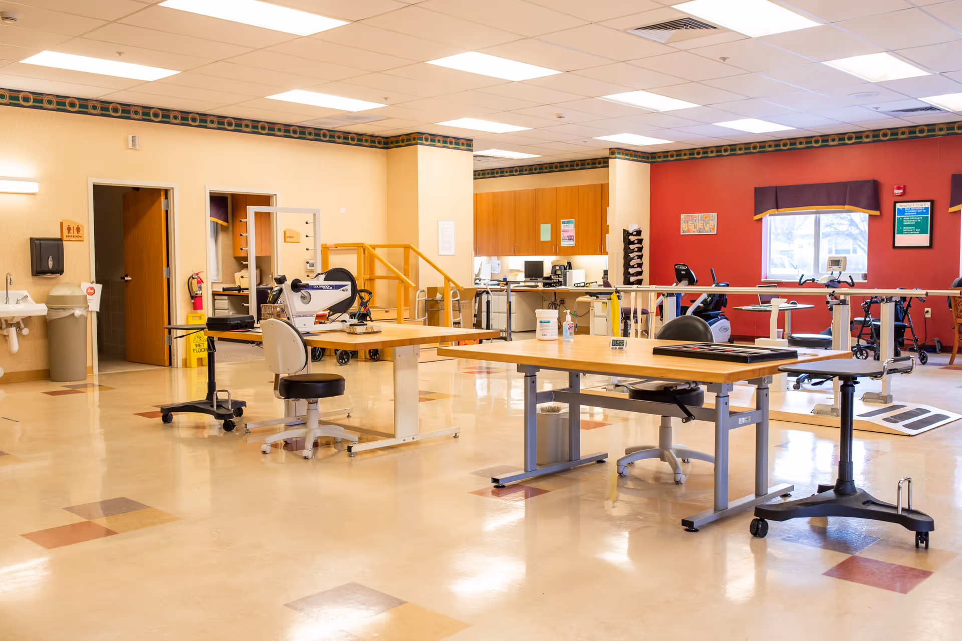A bright rehabilitation room with work tables, chairs, exercise equipment, parallel bars and a small set of stairs.