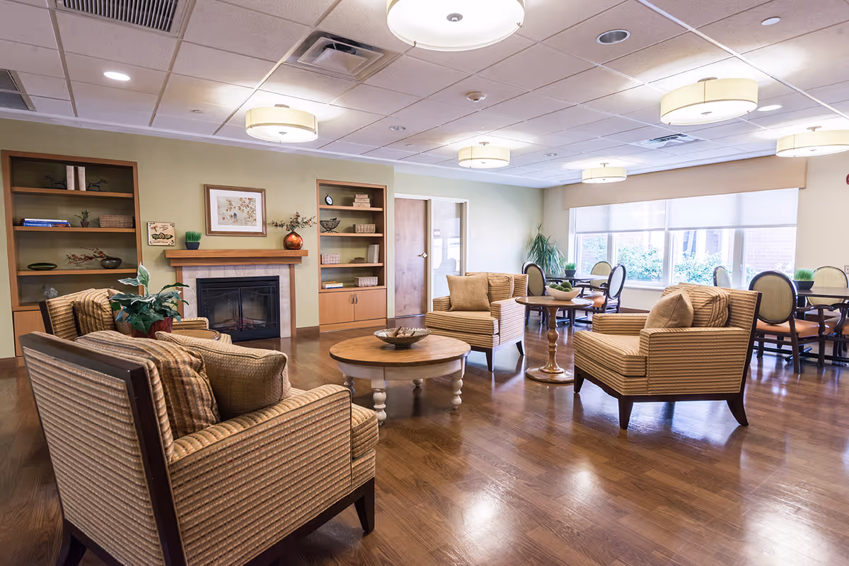 A spacious and well-lit common area with wooden flooring, featuring several cushioned armchairs and a round wooden coffee table in the center. There are built-in wooden shelves on the far wall, a fireplace with a mantle decorated with plants and framed artwork, and large windows letting in natural light. Additional round tables with chairs are visible near the windows.