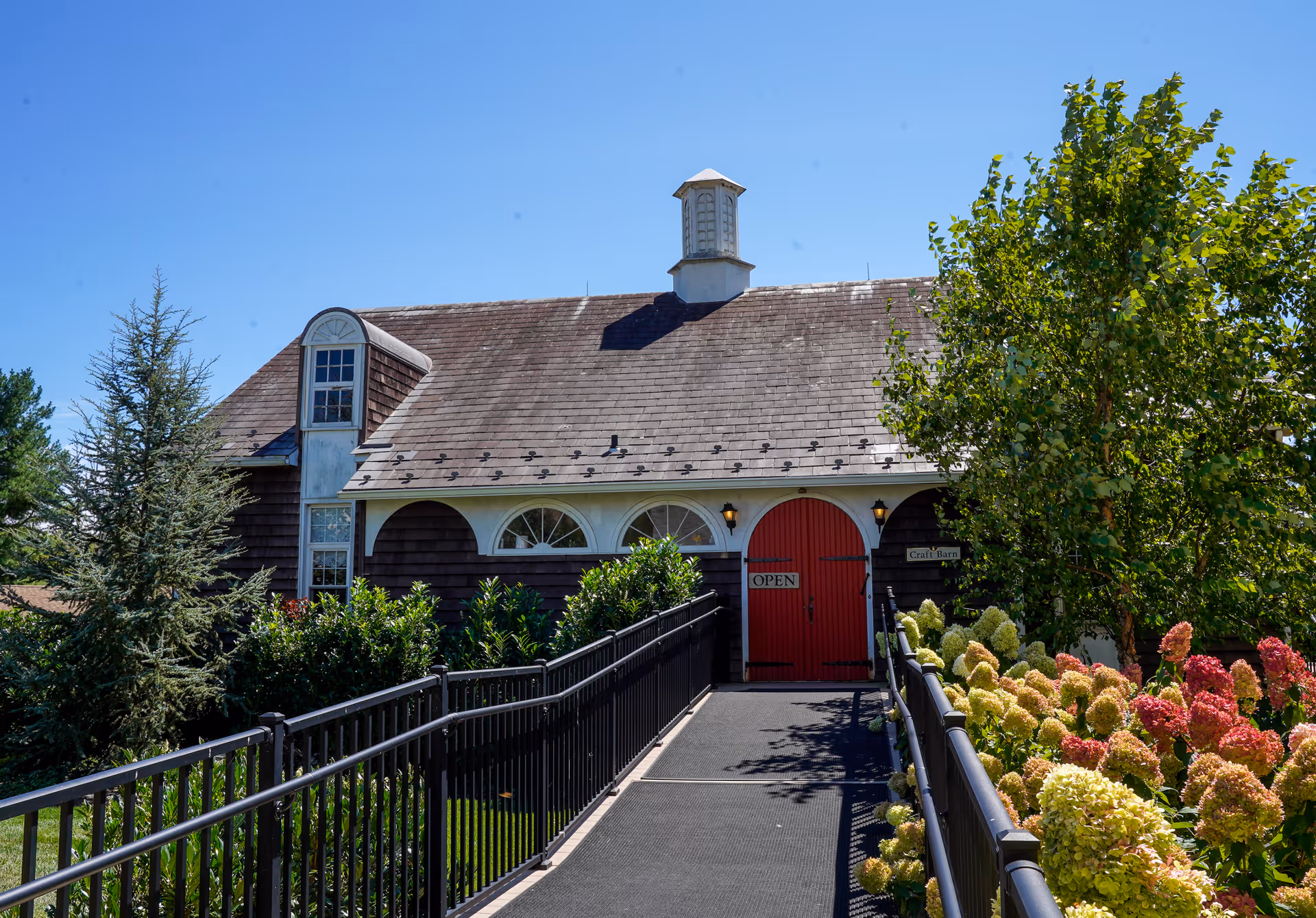 A charming building with a shingled roof and a small cupola on top, featuring a bright red double door with an 'OPEN' sign. The building is surrounded by lush greenery, including flowering bushes and trees, with a black metal railing leading up to the entrance on a sunny day with a clear blue sky.