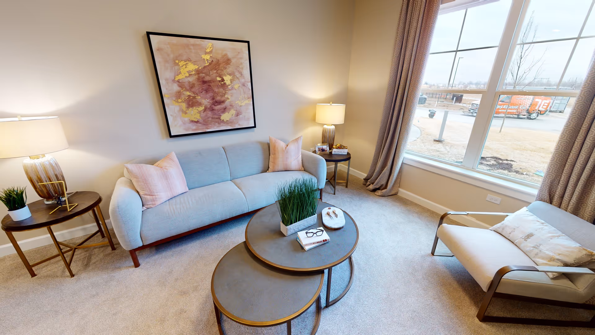 A cozy living room with a light blue sofa adorned with two pink pillows, a modern armchair with a decorative pillow, two round nested coffee tables with a plant and books, two side tables each with a lamp, and a large window with beige curtains letting in natural light. A framed abstract painting hangs on the wall above the sofa.