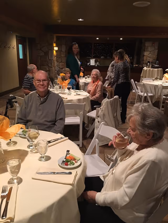 A group of elderly people seated at round tables covered with cream-colored tablecloths in a warmly lit dining room. Some are eating or drinking, while others are standing and conversing. The room has stone walls and wooden beams, creating a cozy atmosphere.