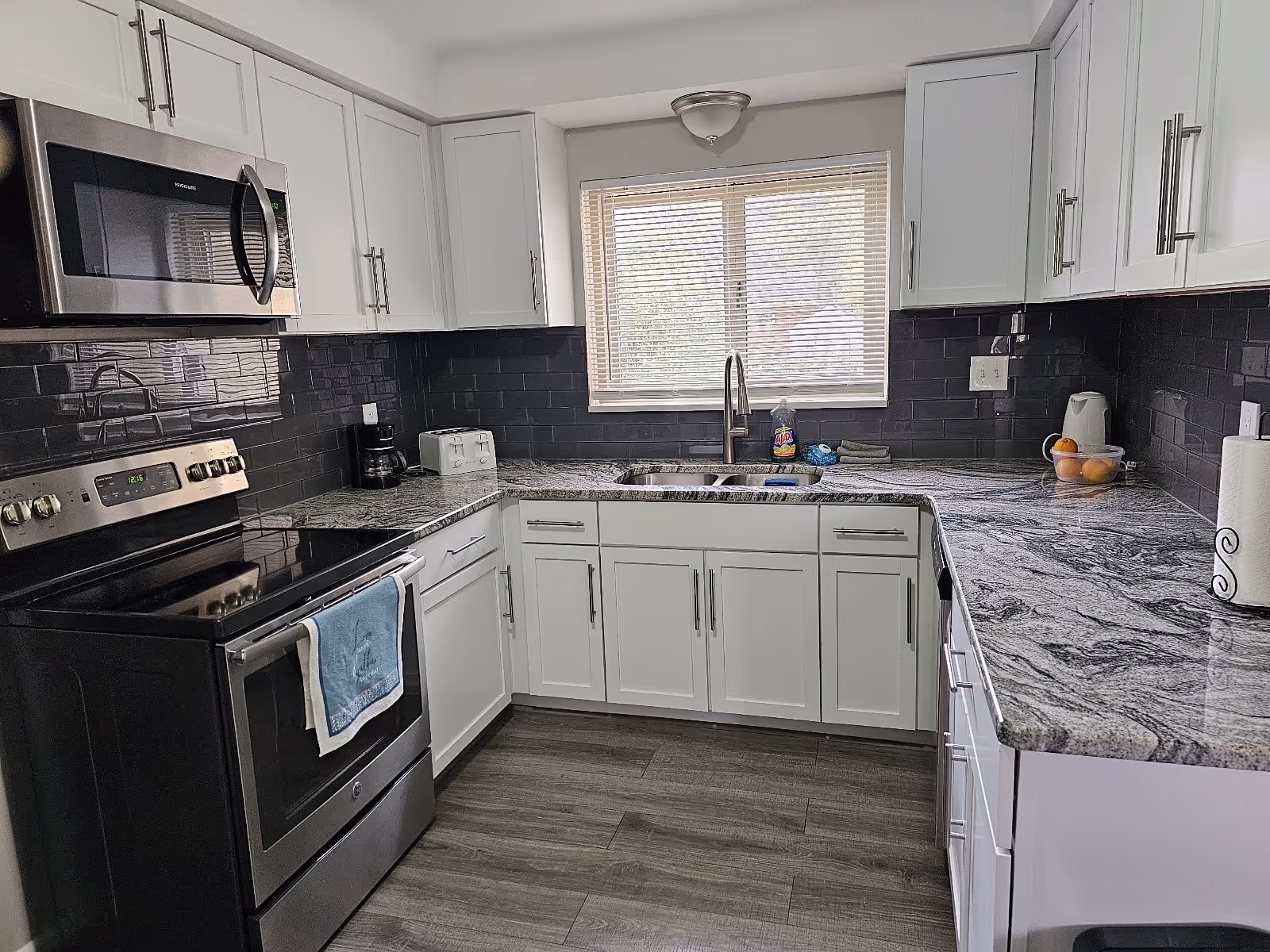 Modern kitchen with white cabinets, gray and white granite countertops, a stainless steel stove and microwave, a double sink under a window with blinds, and various small appliances including a coffee maker, toaster, and electric kettle.