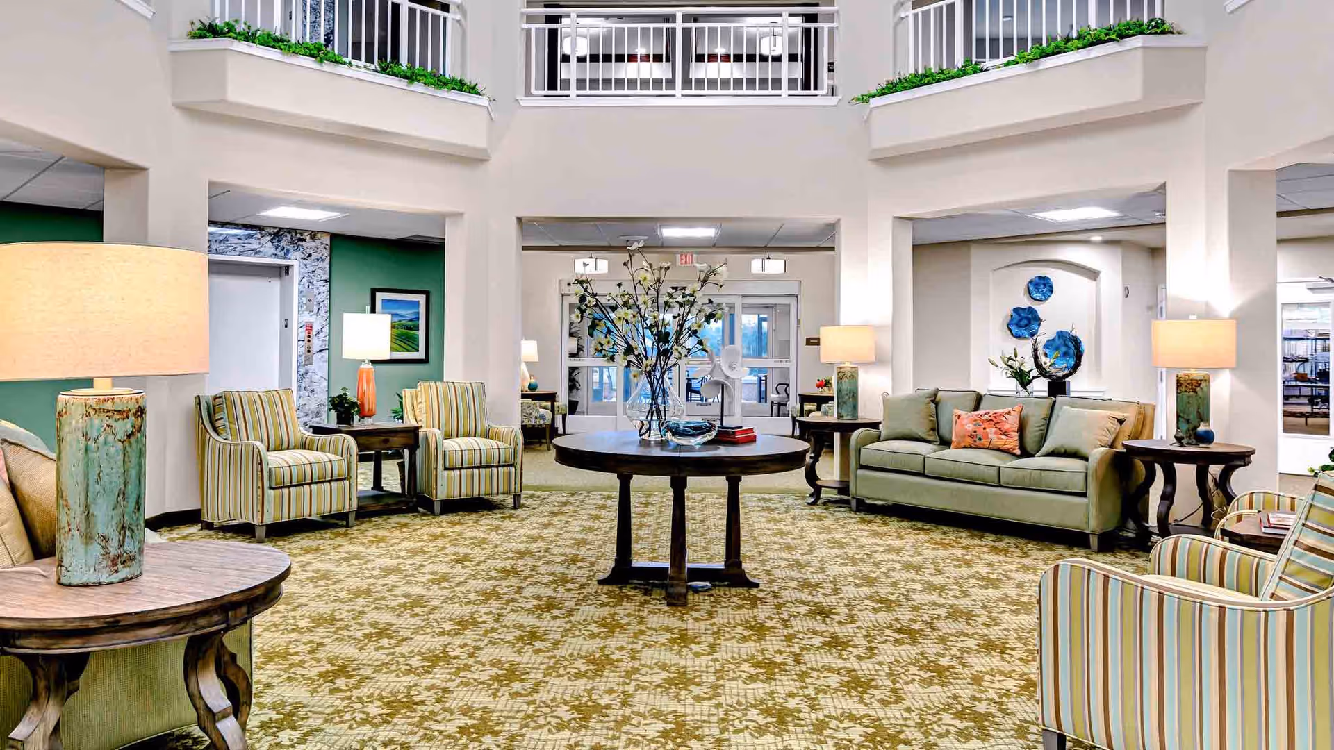 A spacious and well-lit senior living facility lobby with a patterned carpet, green and beige striped armchairs, a green sofa with decorative pillows, wooden side tables with lamps, a round central table with a floral arrangement, and a second-floor balcony with white railings overlooking the area.