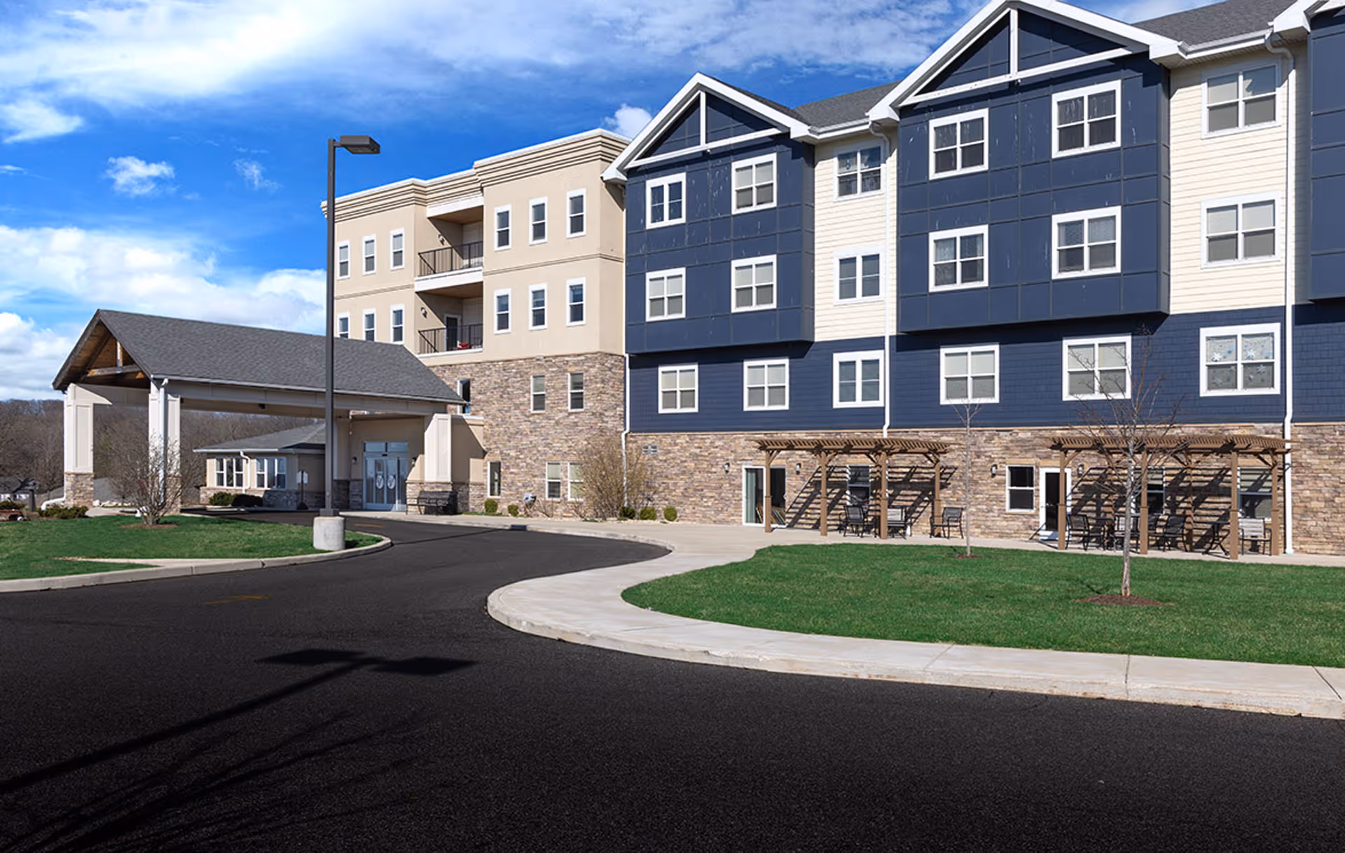 Front exterior of a multi-story senior living building with a covered entrance, curved driveway, lawn, and outdoor seating under pergolas.
