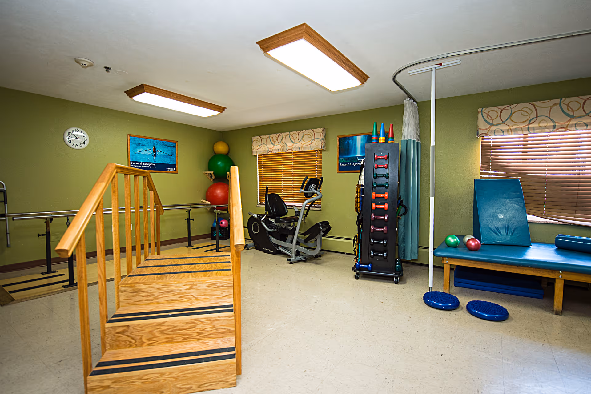 A physical therapy or rehabilitation room with wooden stairs for exercise, parallel bars along the wall, exercise balls, a stationary bike, a rack of colorful dumbbells, and a padded therapy table with exercise equipment. The room has green walls, two windows with blinds and patterned valances, and ceiling lights.