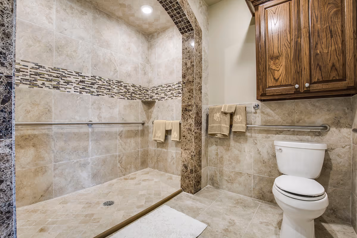 A clean and modern bathroom featuring a walk-in tiled shower with a decorative mosaic tile strip, grab bars for safety, beige towels hanging on the bars, a white toilet, and wooden cabinets mounted on the wall.