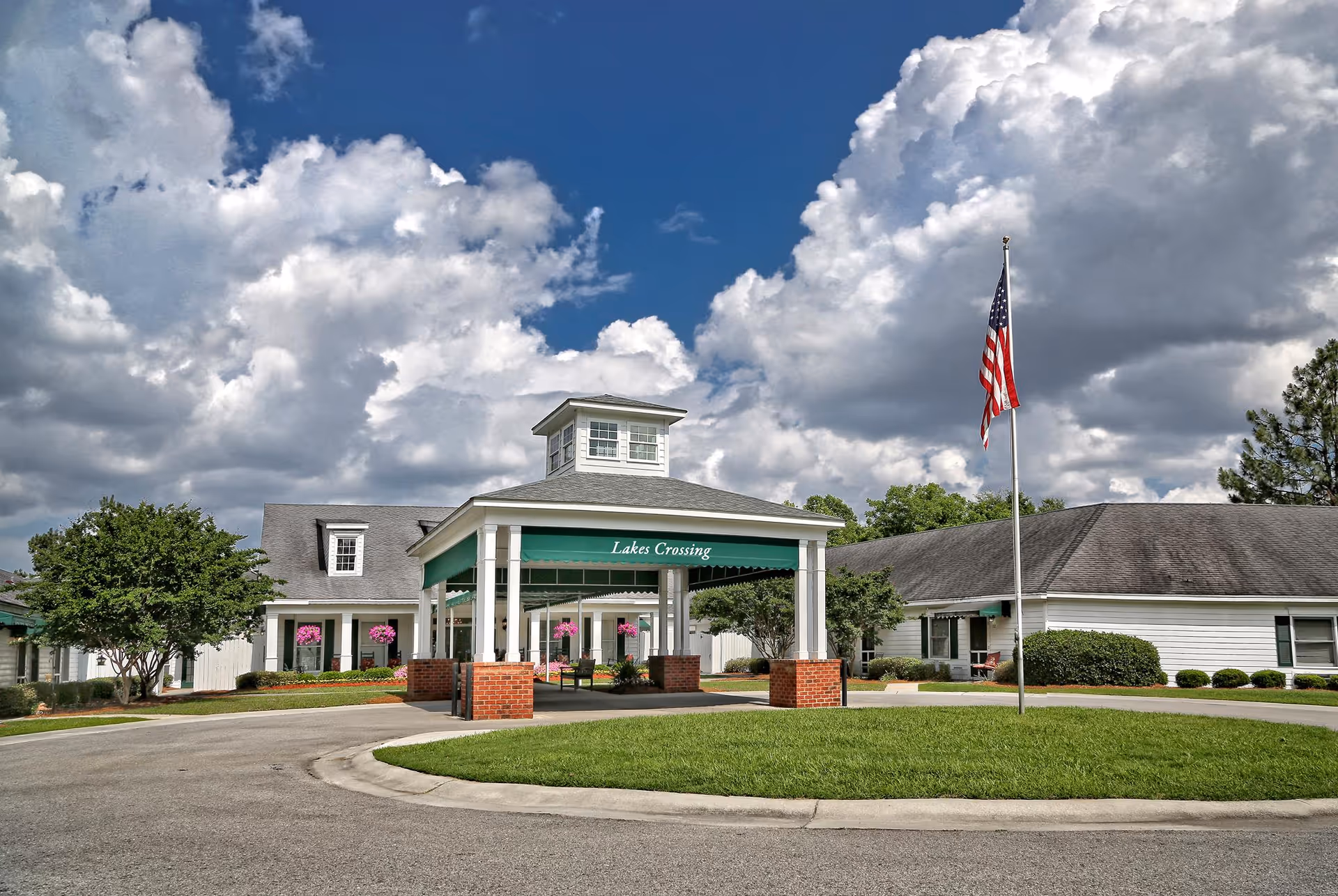 Front entrance of Lakes Crossing Senior Care showing a covered drop-off portico, an American flag on a pole, and the building under a cloudy sky.
