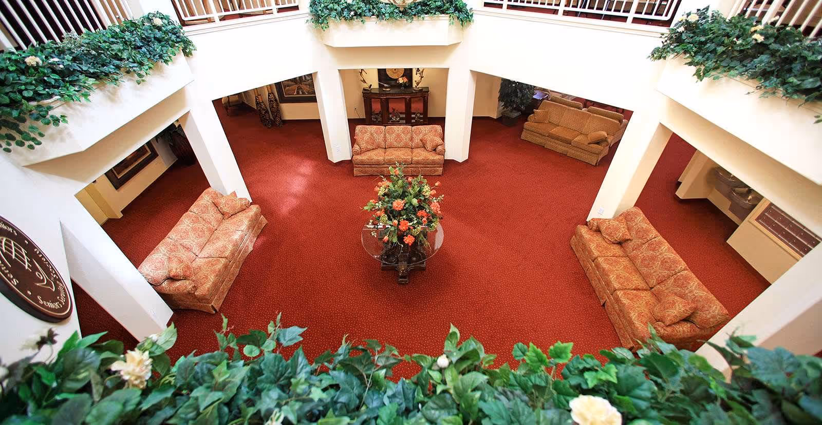 View from an upper level overlooking a spacious living area with red carpet, four patterned sofas arranged around a central glass table with a floral arrangement, white pillars, and greenery along the balcony edges.