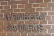 Metallic letters spelling 'WEINBERG GARDENS' mounted on a brick wall.