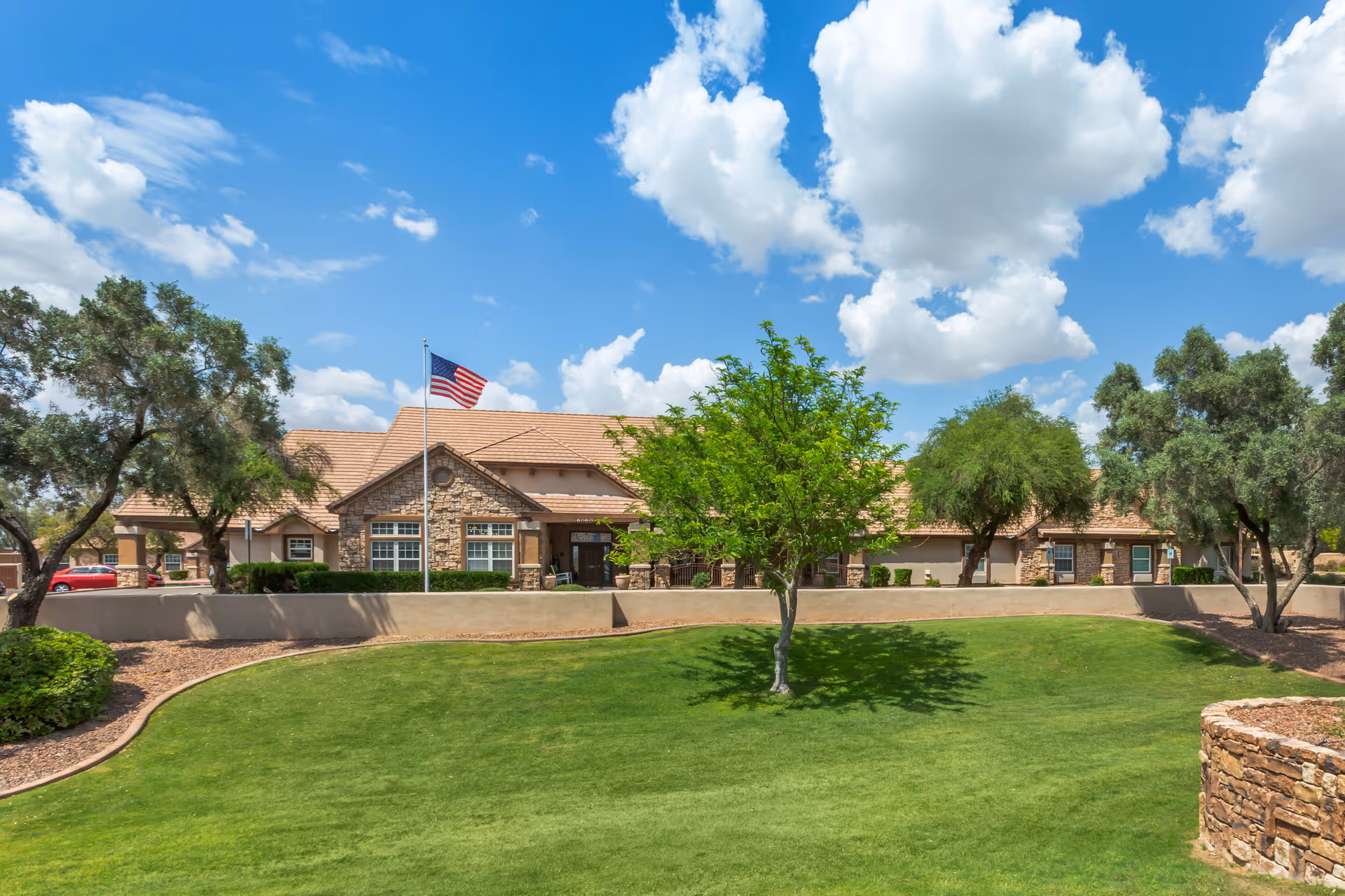 Exterior view of a senior living facility building with a stone facade and tan roof, surrounded by green grass, trees, and a clear blue sky with white clouds. An American flag is flying on a flagpole in front of the building.