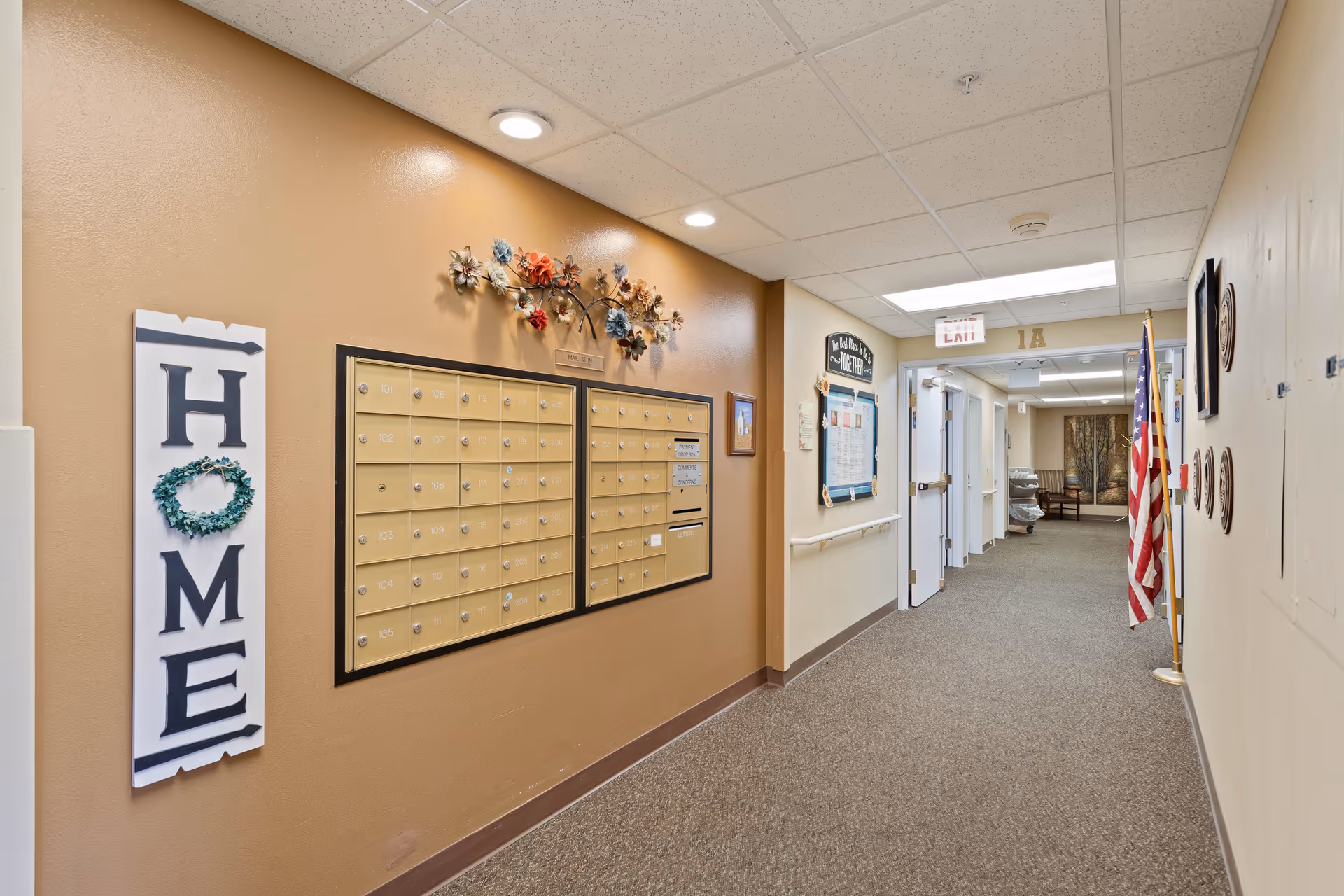 Interior hallway of a senior living facility with a wall-mounted mailbox unit, a decorative sign spelling 'HOME' with a wreath as the letter O, and an American flag near the end of the corridor. The hallway has beige walls, carpeted floor, ceiling lights, and handrails along the walls.
