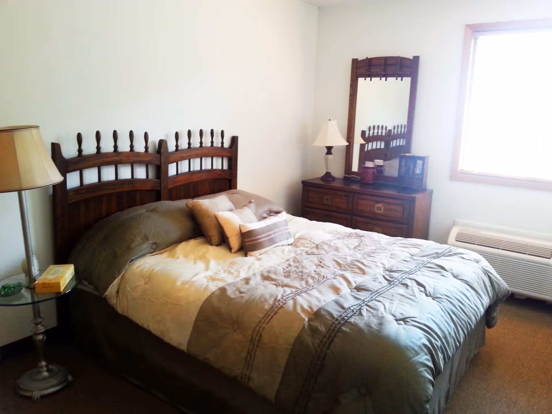 A cozy bedroom featuring a wooden bed with pillows and comforter, a nightstand and lamp, and a dresser with mirror near a window.