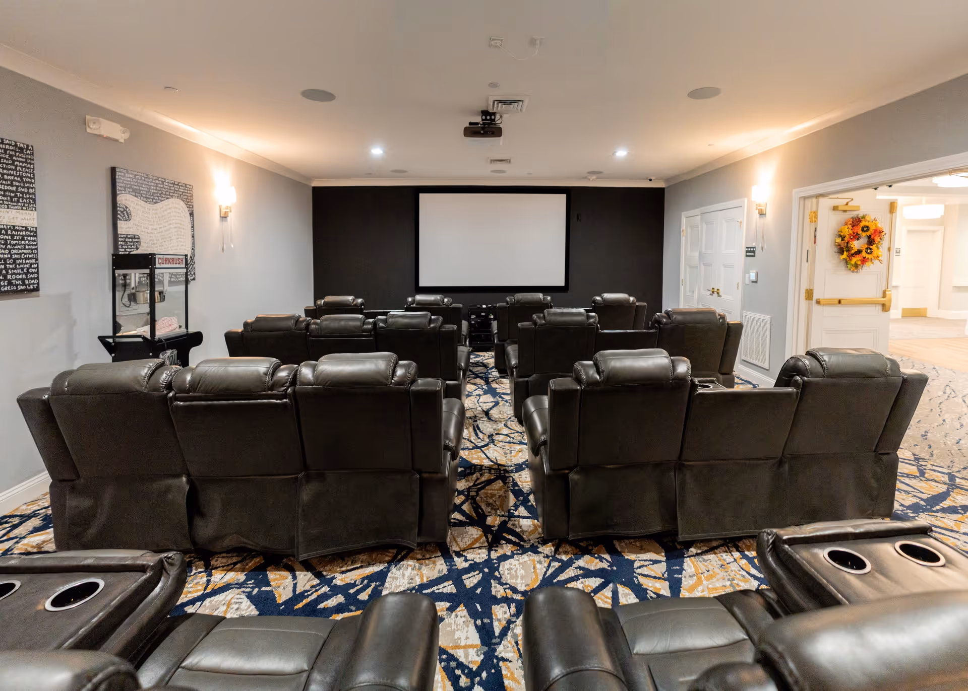 A cozy movie theater room with multiple rows of black leather recliner chairs facing a large blank projector screen on a black accent wall. The room has patterned carpet with blue, beige, and white colors, soft wall lighting, and a popcorn machine on the left side. An open doorway on the right leads to a hallway decorated with a sunflower wreath on the door.