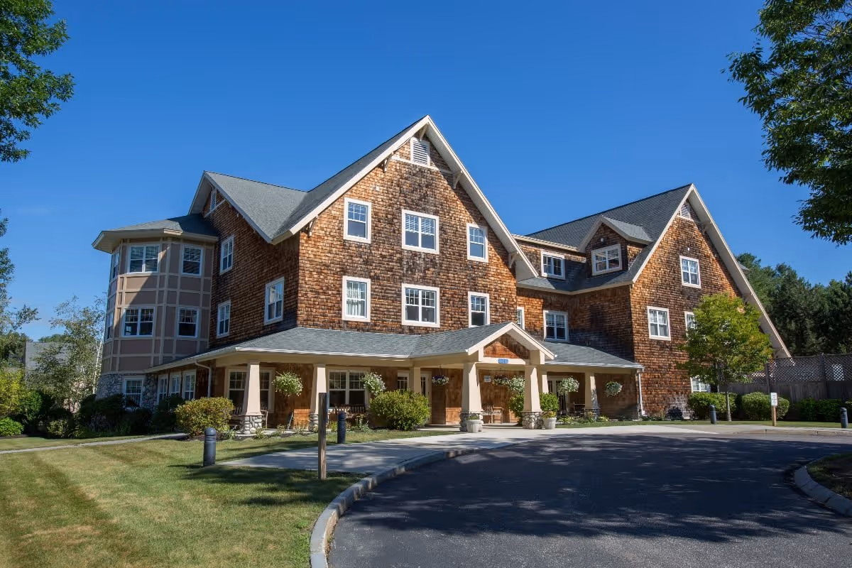 Exterior view of a large, multi-story senior living facility with wooden shingle siding, multiple windows, and a covered entrance. The building is surrounded by well-maintained landscaping, including green grass, bushes, and trees under a clear blue sky.