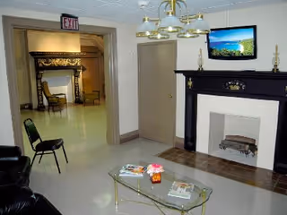 Interior view of a senior living facility lounge area with a black fireplace, a glass coffee table with magazines and a flower vase, a black leather chair, and a doorway leading to another room with chairs and a decorative fireplace. A flat-screen TV is mounted above the fireplace, and a chandelier hangs from the ceiling.