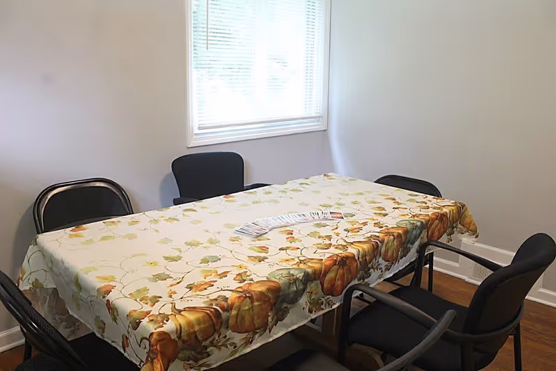 A small dining area with a rectangular table covered by a tablecloth decorated with pumpkins and autumn leaves. Five black chairs surround the table, and a deck of playing cards is spread out in the center. A window with blinds is on the wall behind the table, letting in natural light.
