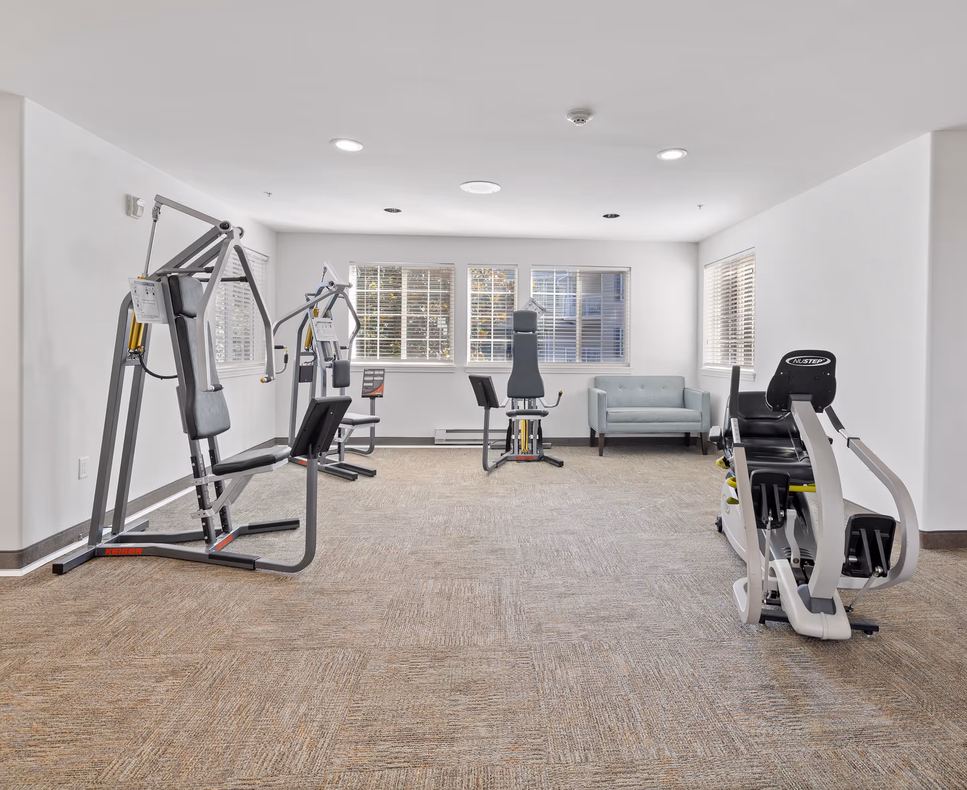 A bright exercise room with several pieces of gym equipment including a weight machine and a seated leg press. The room has beige carpet, white walls, and three large windows with blinds letting in natural light. A light blue loveseat is placed against the far wall.