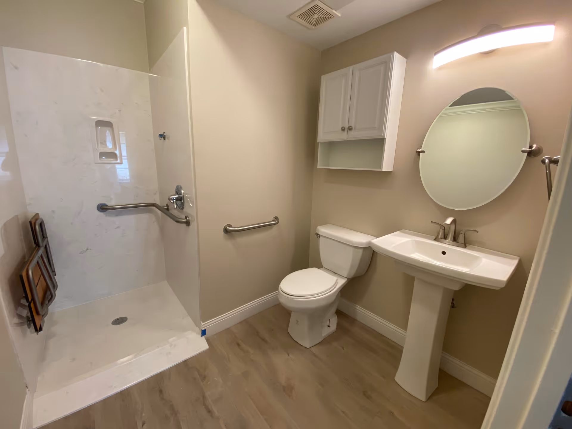 A clean and accessible bathroom featuring a walk-in shower with grab bars, a white toilet, a pedestal sink with a modern faucet, an oval mirror above the sink, a wall-mounted cabinet, and light wood flooring.