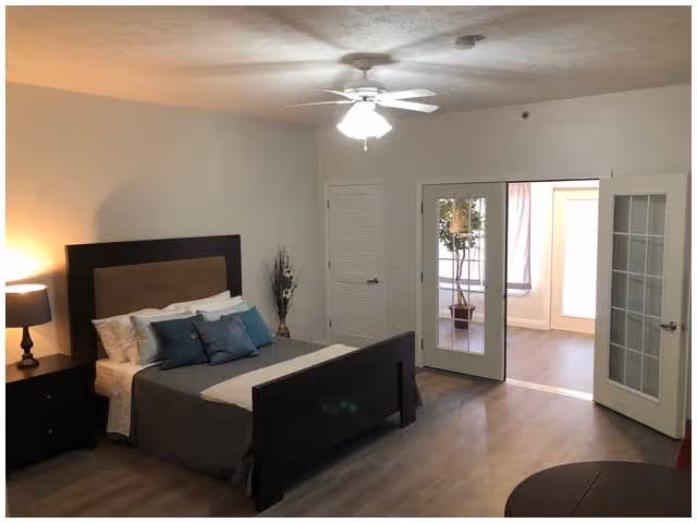A bedroom with a dark wooden bed frame and headboard, neatly made with white and gray bedding and blue accent pillows. There is a nightstand with a lamp on the left side of the bed, a ceiling fan with a light above, and a vase with decorative branches in the corner. Double glass doors open to another room with a potted plant visible inside. The floor is wood, and the walls are painted white.