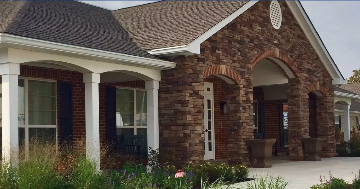 Exterior view of a senior living facility with a brick and stone facade, white columns, large windows, and a covered entrance with arches. There are plants and flowers in the foreground.