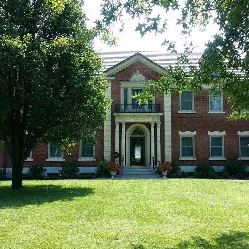Front exterior view of a two-story brick building with white trim and columns at the entrance. The building is partially shaded by large green trees and has a well-maintained green lawn in front. There are flower pots with red flowers on either side of the entrance steps.