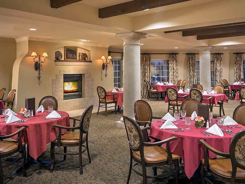 Dining room with round tables covered in red tablecloths, set with white napkins, glassware, and floral centerpieces. The room features upholstered chairs, a stone fireplace with a fire burning, wall sconces with lamps, and large windows with patterned curtains.
