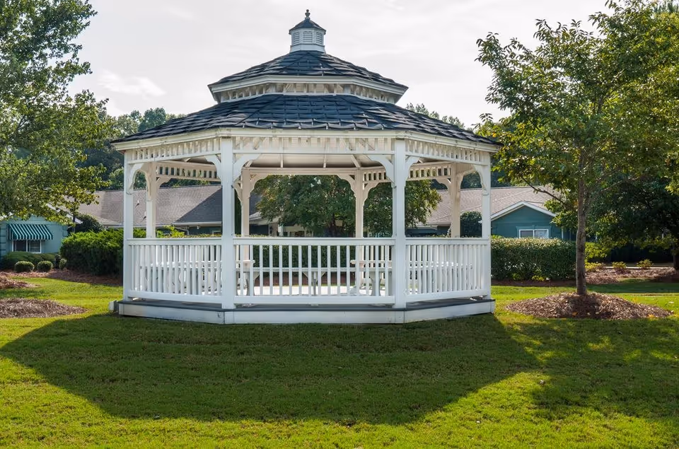 A white wooden gazebo with a shingled roof situated on a well-maintained grassy lawn, surrounded by trees and shrubs, with residential buildings visible in the background under a partly cloudy sky.