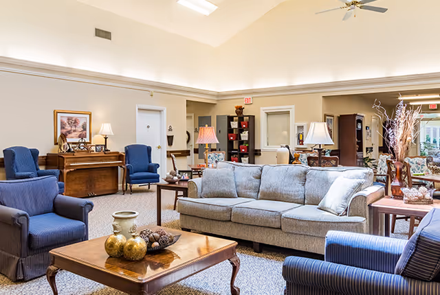 A spacious and well-lit living room area in a senior living facility with comfortable seating including a gray sofa and blue armchairs arranged around a wooden coffee table. The room features a piano against the wall, decorative lamps, framed artwork, and various decorative items on tables and shelves. The ceiling is high with recessed lighting and a ceiling fan.