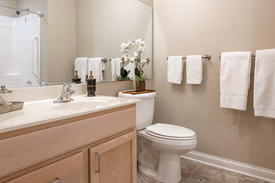 Clean, neutral-toned bathroom with a sink vanity, large mirror, toilet, towel racks, and a shower in the background.
