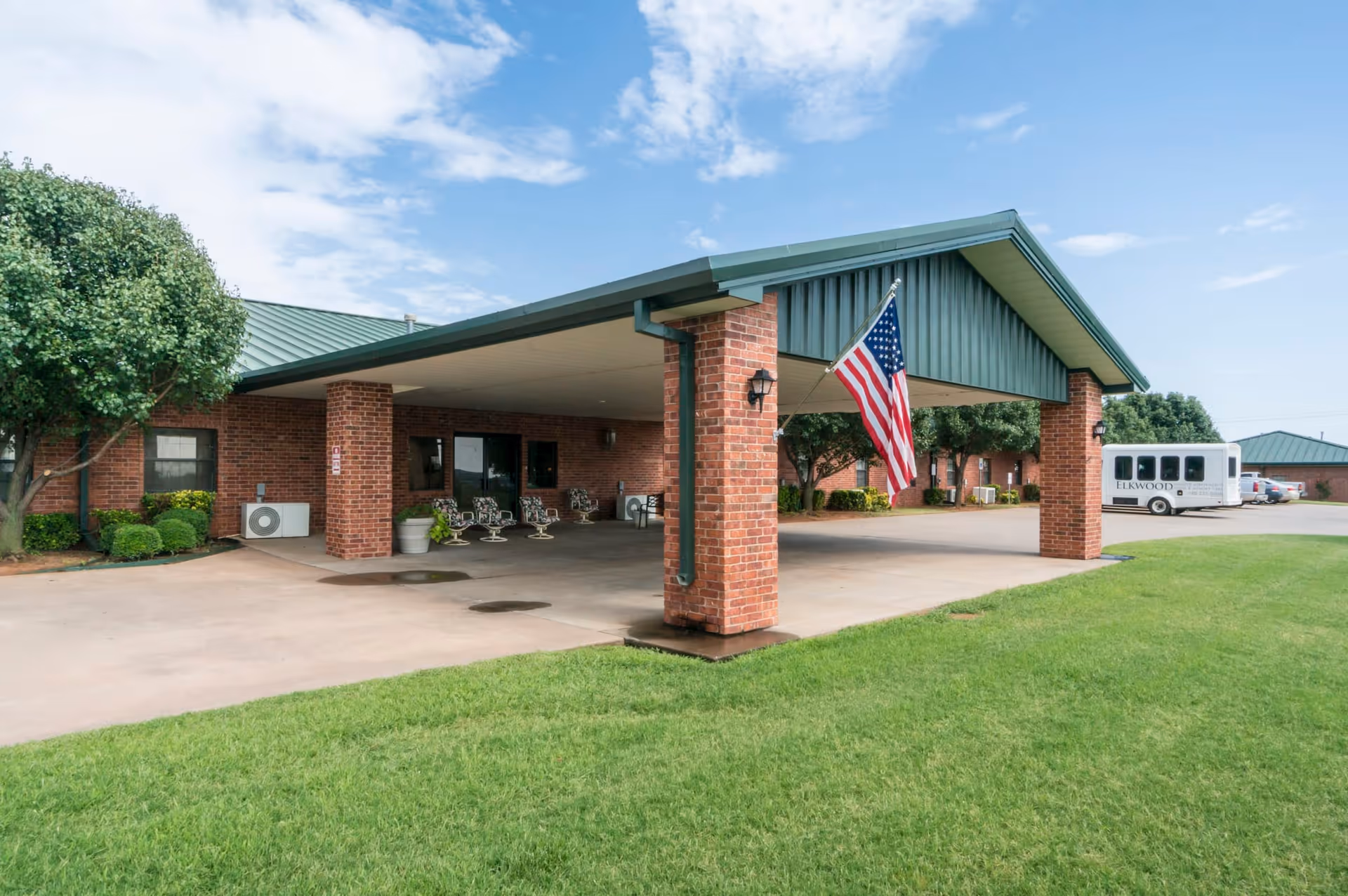 Exterior view of a senior living facility entrance with a covered drop-off area supported by brick pillars. An American flag is hanging from one of the pillars. There are several chairs under the covered area, green grass in the foreground, and a white shuttle bus parked in the background.