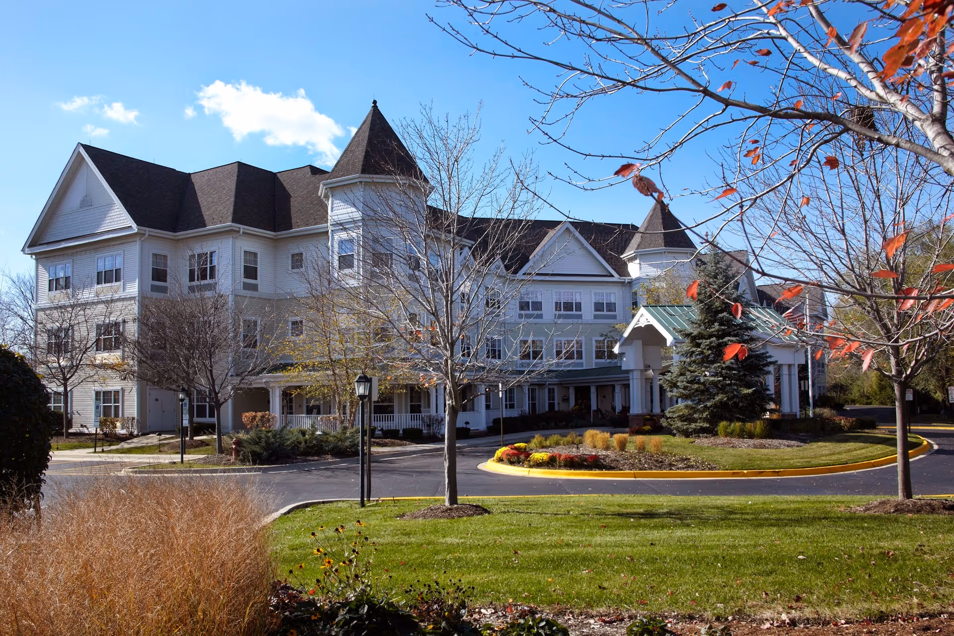Exterior view of a large, white multi-story senior living facility with a turret-like architectural feature, surrounded by landscaped grounds with trees, shrubs, and a circular driveway under a clear blue sky.